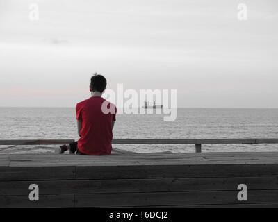Junge Menschen allein an der Promenade am Schiff suchen sitzen vor der Küste verankert, Musik hören, (Konzept der Einsamkeit, Entspannung). Stockfoto