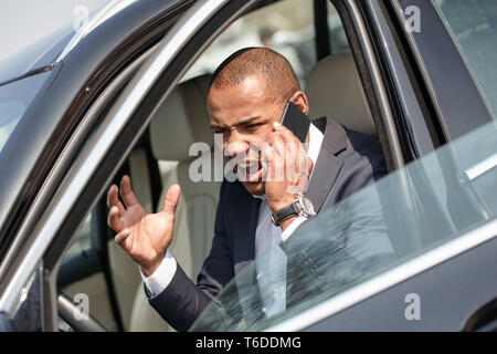 Junge afrikanische amerikanische Geschäftsmann sitzen im Auto Tür geöffnet halten Smartphone argumentieren mit Anrufer wütend Vorderansicht Stockfoto