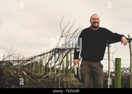 Der bärtige Mann mit Axt und Entenschnabel neben einem neu erbauten traditionellen Hedge stehend, Lachen. Stockfoto