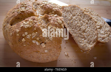 Ein runder Laib artisan Vollkornbrot mit zählte Kruste, garniert mit Haferflocken, Nahaufnahme, ruht auf einem Bambus Schneidebrett. Stockfoto