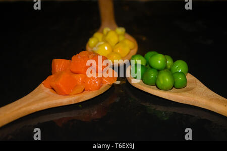 Stücke von Karotten, mais und Erbsen auf Holzlöffel mit dunklen Hintergrund Stockfoto