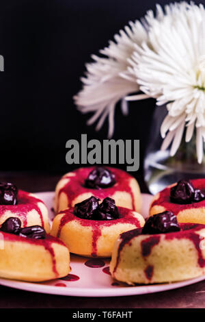 Sauerkirschen und Ziegenkäse Donuts auf einem Rosa Platte vor einem schwarzen Hintergrund mit weißen Blumen in einer Vase hinter Stockfoto