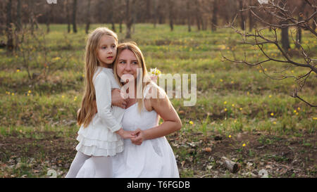 Family Portrait einer alleinstehenden Mutter ihre Tochter umarmte. Sie posiert in weißen Kleidern vor dem Hintergrund einer Frühlingswiese. Muttertag. Die gi Stockfoto