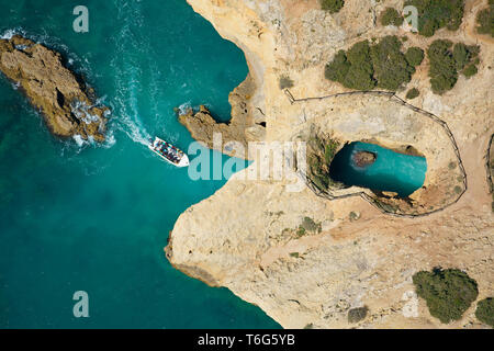 LUFTAUFNAHME. Sightseeing-Boot nähert sich einer Meereshöhle / Sinkhole an der felsigen Küste in der Nähe von Cão Raivoso Beach. Lagoa, Algarve, Portugal. Stockfoto