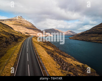 Luftaufnahme von einer Küstenstrasse entlang einer Färöischen Fjord mit atemberaubenden Bergketten, dramatischer Himmel und türkisfarbenem Wasser (Färöer, Dänemark, Europa) Stockfoto
