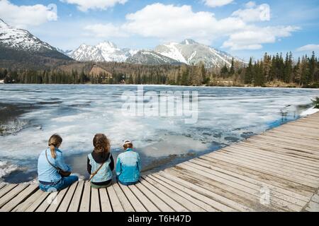 Rückansicht auf drei weibliche Touristen sitzen durch gefrorene Bergen See Strbske Pleso - Frühling in der Hohen Tatra in der Slowakei. Stockfoto