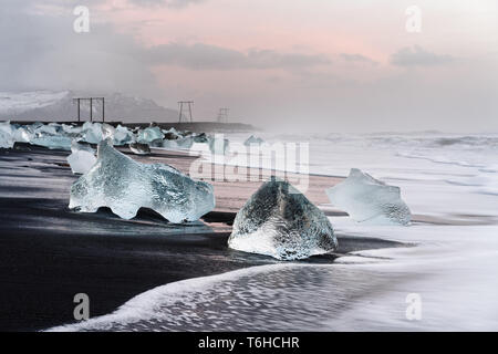 Eisblöcke sind malerisch auf einem schwarzen vulkanischen Strand, die Bewegung der Wellen gesehen werden kann (langzeitbelichtung), darüber ein bewölkter Himmel mit Stockfoto