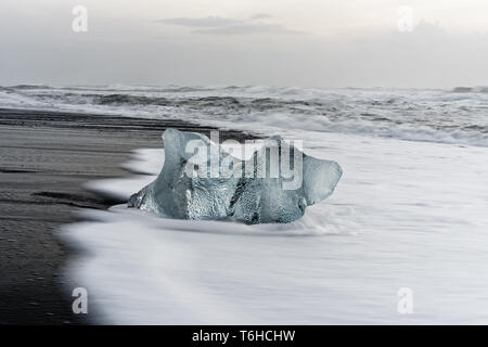 Markante Eisblock in blauen Farbtönen auf einem schwarzen Strand mit starker Brandung, Gischt auf der dunklen Sand, Wellen im Hintergrund - Ort: Island, Jöku Stockfoto