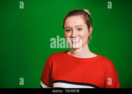 Eine Frontansicht Portraitfotos eines jungen kaukasischen stehende Frau vor einem Green Screen, sie lächelt und schaut in die Kamera. Stockfoto