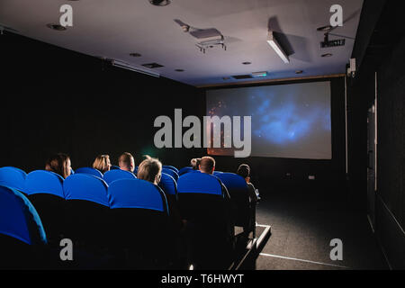 Eine Ansicht von hinten erschossen von einem Film Theater, unerkennbare Personen sitzen zusammen in blau Sitze in einer Reihe vor der Leinwand gesehen werden kann. Stockfoto