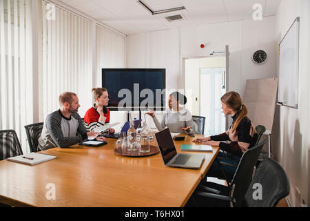 Einen weiten Blick Schuß eines kleinen multi-ethnischen Gruppe von Menschen an einem Tisch im Büro, in der Menschen gesehen werden kann über die Arbeit mit ihren Kollegen Stockfoto