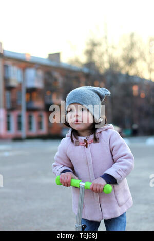 Süße Mädchen in einen Hut, Mantel und Jeans, einem Dreirädrigen Roller. Abend Frühling Stadt Sonnenuntergang. Stockfoto