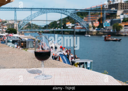 Selektiver Fokus Rotwein Glas mit Blick auf Cais da Ribeira und Ponte de Dom Luis I auf dem Douro in Porto, Portugal Stockfoto