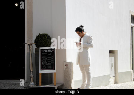Eine schöne elegante junge Frau auf der Suche bei Mobile in modischen weißen Hosen Anzug Mode in Alfama Straße vor dem Restaurant Lissabon Portugal KATHY DEWITT Stockfoto