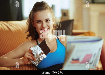 Lächelnd aktive Frau in der Fitness Kleidung in das moderne Haus lesen Magazin und Schokolade essen. Stockfoto