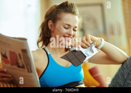 Gerne sportlich aktive Frau in Fitness Kleidung in das moderne Haus lesen Magazin und Schokolade essen. Stockfoto