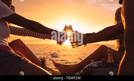 7/8 Schuß von Freunden in der Nähe von dem Meer mit Speisen und Getränken bei Sonnenuntergang sitzen. Fröhliche Freunde auf Urlaub toasten Getränke sitzen am Strand mit Sonne in Stockfoto