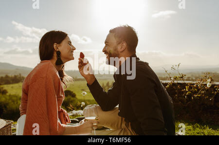 Glückliches Paar auf einem Datum sitzen in Wine Farm reden und Spaß haben. Mann Fütterung eine Erdbeere zu seiner Freundin in einem Weinberg sitzen an einem sonnigen Tag. Stockfoto