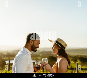 Glückliches Paar an jedem anderen Holding Glas Rotwein in einem Weingut. Seitliche Sicht auf ein lächelndes Paar in Liebe miteinander sprechen, Toasten von Glas Stockfoto