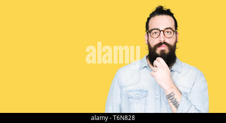 Junge hipster Mann mit langen Haaren und Bart Tragen einer Brille in die Kamera schaut zuversichtlich mit Smile mit verschränkten Armen und Hand am Kinn angehoben. Denken Stockfoto