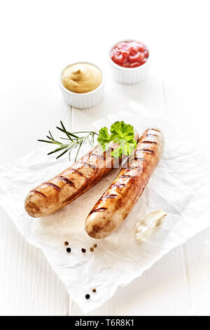 Teil Zwei gegrillte Würstchen serviert auf weisse Serviette mit Gewürzen und frische Petersilie auf hölzernen weissen Tisch. Ketchup und Senf, serviert in kleinen weißen Stockfoto