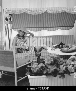 Sommer der 60er Jahre. Eine junge Frau sitzt in einer Hängematte comforably trinken Kaffee. Das Design der Möbel und ihr Kleid stellt die 1960er Dekade sehr gut. Schweden 1960 Stockfoto