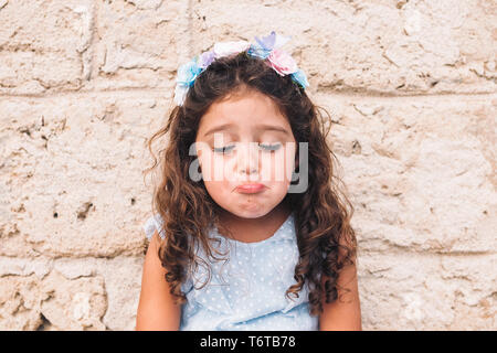 Kleines Mädchen Spaß, mit einem traurigen Gesicht, ist vor einer Mauer aus Stein und trägt ein blaues Kleid und eine Blume Kopfbügel Stockfoto