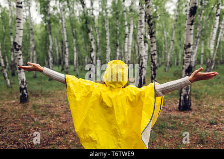 Glückliche junge Frau im gelben Regenmantel Wandern im Frühjahr Wald unter Regen und Spaß Anheben der Arme. Das Leben genießen Stockfoto