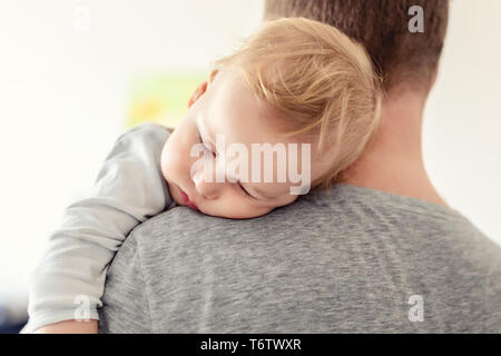 Portrait von cute adorable Blonde kaukasischen Toddler boy Schlafen auf Väter Schulter im Innenbereich. Süßes kleines Kind das Gefühl von Sicherheit und Versorgung auf Daddy's Hand. Stockfoto