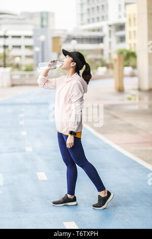 Eine junge und aktive asiatische Frau runner ist das trinken eine Flasche Wasser für Erfrischungen nach dem Ausführen im blauen Laufbahn in einem Park Stockfoto