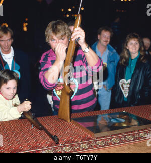 Der deutsche Fernsehmoderator, Entertainer, Wis, Hörbuchsprecher, Musiker und Komiker Jürgen von der Lippe eine Einems Schießstand auf einer Kirmes, Deutschland 1980er. Der Deutsche Fernsehmoderator, Entertainer, Schauspieler, Sprecher, Musiker und Komiker Jürgen von der Lippe zu einem Shooting Gallery an Karneval, Deutschland der 80er Jahre. Stockfoto