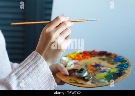 In der Nähe der weiblichen Händen in einem rosa Pullover mit einem Besen und eine Palette mit bunten Acrylfarben gegen eine weiße Leinwand, selektiven Fokus Stockfoto
