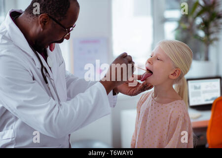 Dunkelhäutige Kinderarzt gefragt, kleines Mädchen Mund zu weit geöffnet Stockfoto