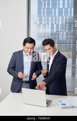 Seitenansicht von zwei schweren junge Geschäftsleute, die in schwarzen und blauen Anzügen Kaffee trinken und reden in der Nähe großer Fenster zuversichtlich Partner diskutieren ide Stockfoto