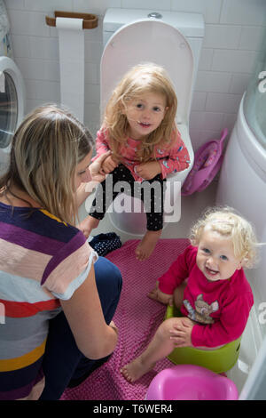 Junge Kinder in der Toilette mit ihrer Mutter, einer auf der Toilette sitzt, während ihre jüngere Schwester verwendet ein Töpfchen. Stockfoto