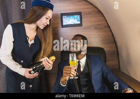 Junge erfolgreiche afro-amerikanischen Geschäftsmann, der in Gläsern und einer stewardess zeigt eine Flasche Wein in der Kabine eines privaten Jet. Service und Fliegen erste Stockfoto