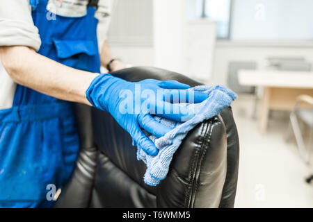 Junger Mann in Arbeitskleidung und Gummihandschuhe reinigt den Bürostuhl mit professioneller Ausstattung. Stockfoto