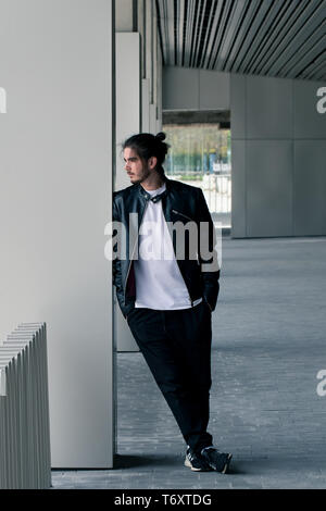Bärtige hübscher junger Mann in eine schwarze Lederjacke mit einem stilvollen Haarschnitt steht und lehnt sich an der Wand, auf dem Weg schauen. Stockfoto