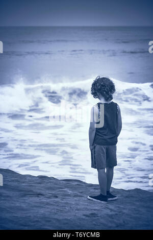 Ein Junge mit dem lockigen Haar zu tragen Tanktop und Shorts steht am Strand und blickt auf das Meer an bewölkten Tag in San Diego, Kalifornien. Stockfoto