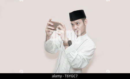 Portrait von Stattlichen asiatischen muslimischen Mann mit Kopf und mit Smartphone und ernsthaft zu spielen video game, muslimische auf e Sport Konzept - Bild Stockfoto