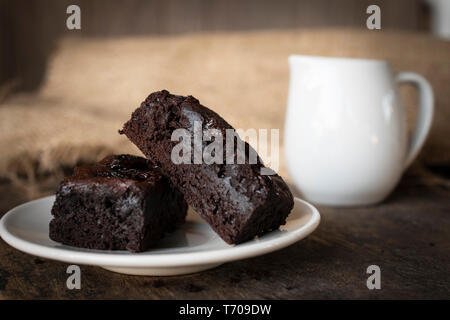 Brownie Kuchen auf einem weißen Teller mit einem Glas weiße Milch daneben platziert. Das Setzen auf einen Schreibtisch aus Holz. Stockfoto