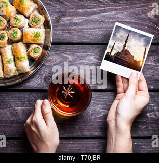 Hände mit Foto von der Blauen Moschee in Istanbul und schwarzer Tee in der Nähe der Türkischen baklava auf hölzernen Hintergrund Stockfoto