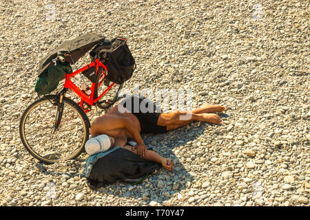 Älterer Mann im Schatten von seinem Fahrrad liegen auf einem Kiesel- und Felsstrand in Alicante Spanien Stockfoto