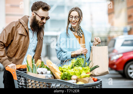 Junge und glückliche Paar gehen mit shoopping Warenkorb voller gesunder Speisen auf dem Parkplatz in der Nähe der Supermarkt Stockfoto