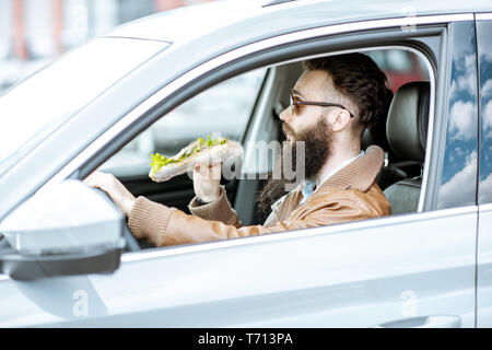 Stilvolle bärtiger Mann einen Snack mit leckeren Sandwich beim Autofahren in der Stadt Stockfoto