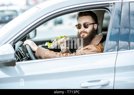 Stilvolle bärtiger Mann einen Snack mit leckeren Sandwich beim Autofahren in der Stadt Stockfoto