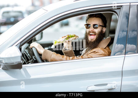 Stilvolle bärtiger Mann einen Snack mit leckeren Sandwich beim Autofahren in der Stadt Stockfoto