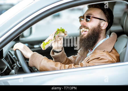 Stilvolle bärtiger Mann einen Snack mit leckeren Sandwich beim Autofahren in der Stadt Stockfoto