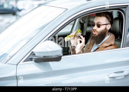 Stilvolle bärtiger Mann einen Snack mit leckeren Sandwich beim Autofahren in der Stadt Stockfoto