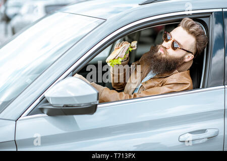 Stilvolle bärtiger Mann einen Snack mit leckeren Sandwich beim Autofahren in der Stadt Stockfoto